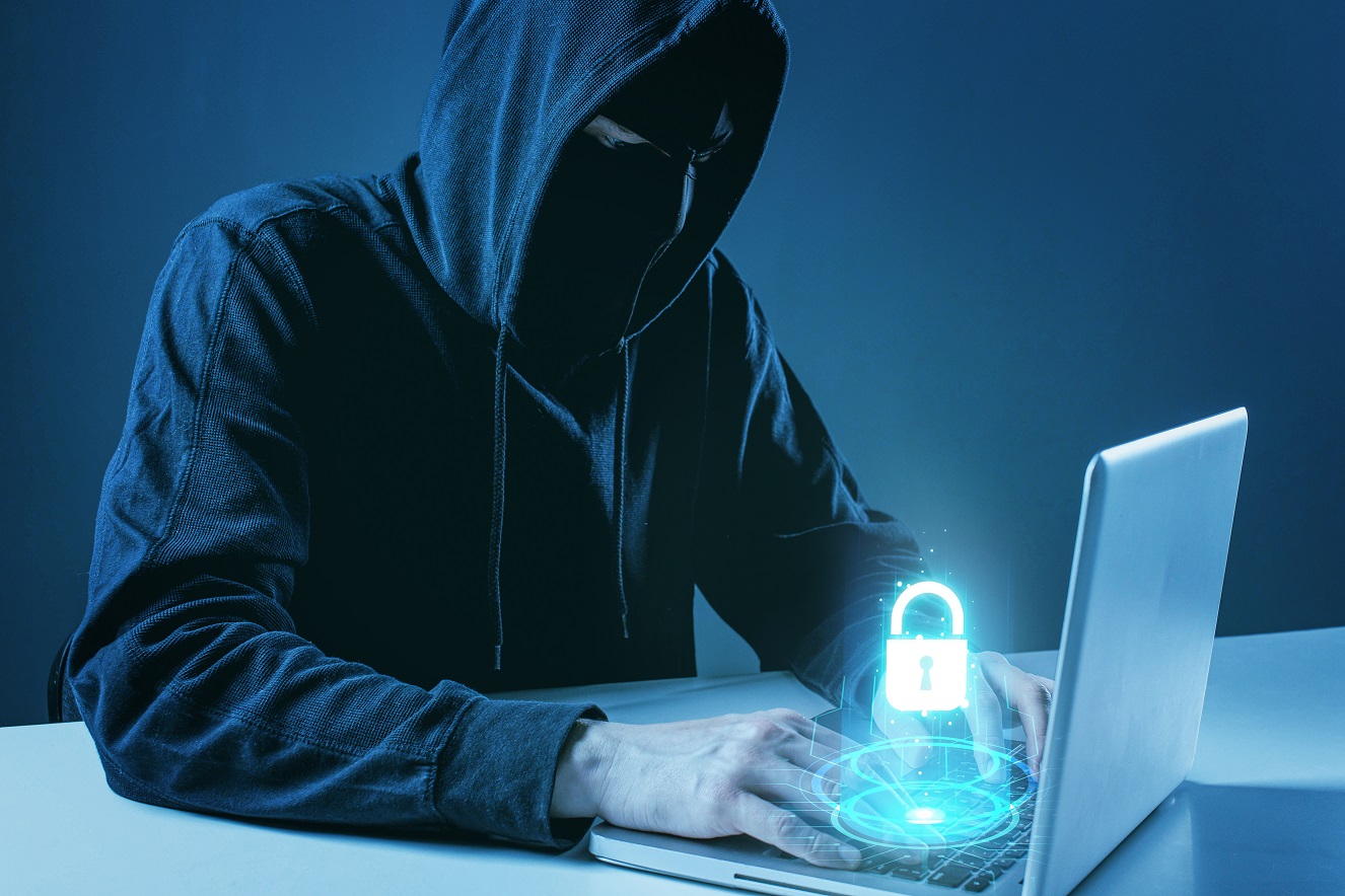 Zamaskowany mężczyzna pisze na klawiaturze laptopa, nad jego palcami unosi się zamknięta kłódka. Pobrano z freepik.com