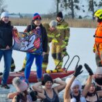 Grupa osób morsuje w wodzie, wokół nich na lodzie strażacy oraz 2 kobiety z flagą