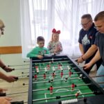 Cztery dorosłe osoby grają przy stole do piłkarzyków. przygląda się dwójka dzieci
